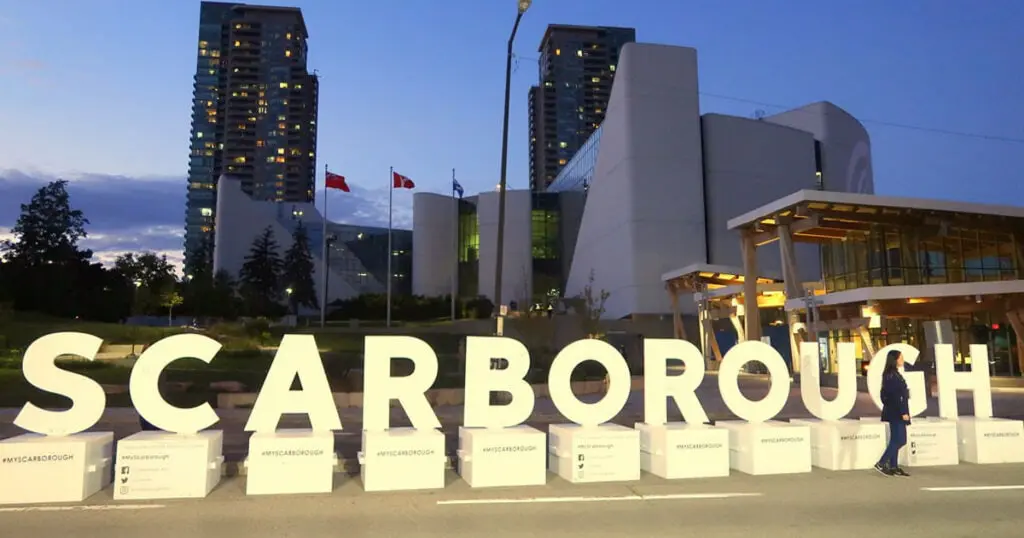 Night view of Scarborough sign with modern buildings and flags in the background, illuminated by city lights.