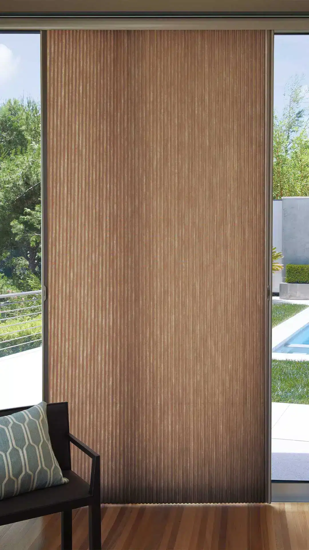 Close-up of a contemporary wooden sliding door with vertical grooved slats, installed inside a bright home with large windows revealing lush outdoor greenery and a pool area.