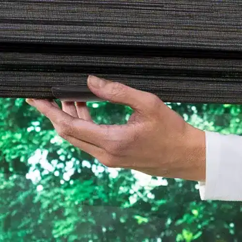 A hand adjusting black horizontal window blinds outdoors with green foliage in the background.