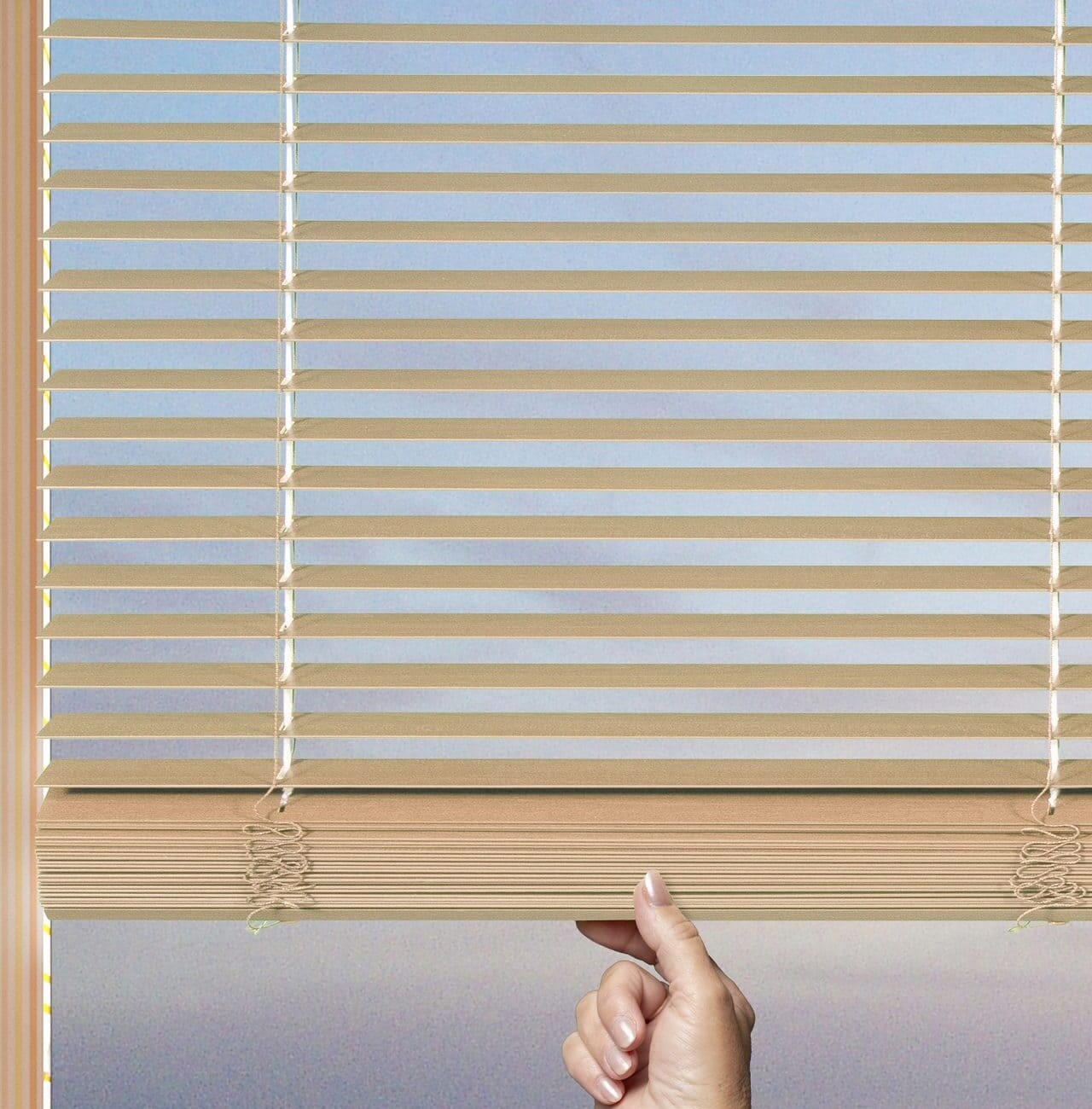 A person’s hand is pulling down the beige window blinds, allowing light and a scenic view to enter the room.
