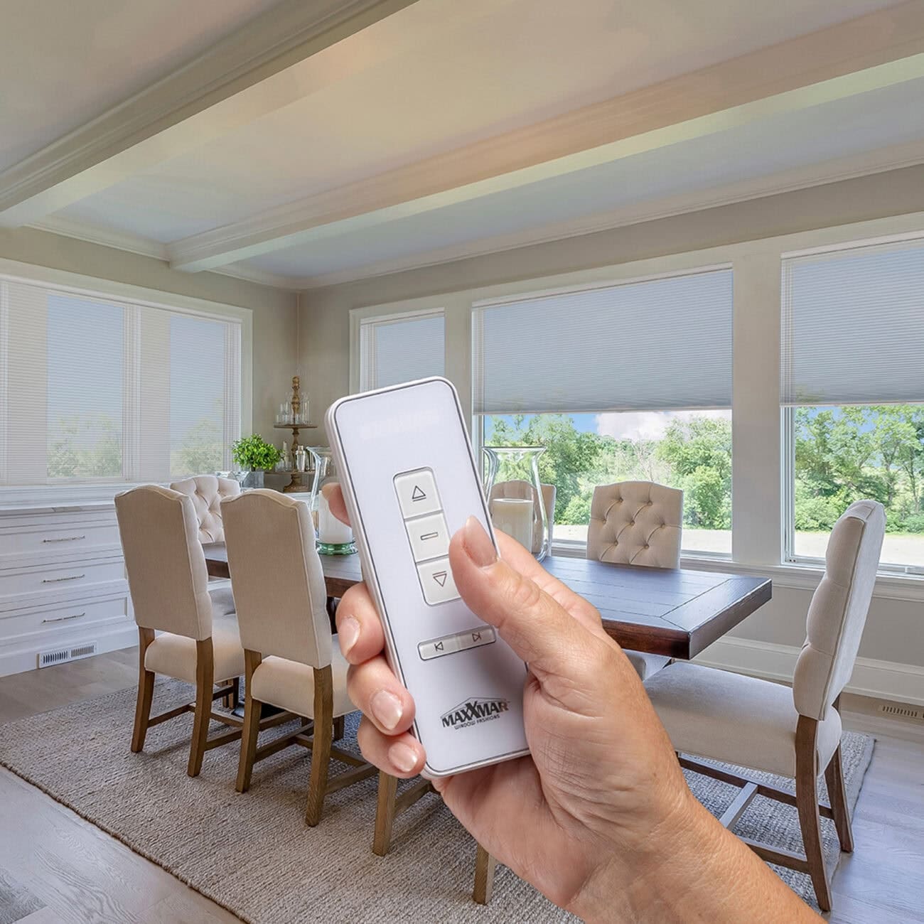Close-up of hand holding remote control for motorized cellular shades in bright dining room with large windows and greenery outside.