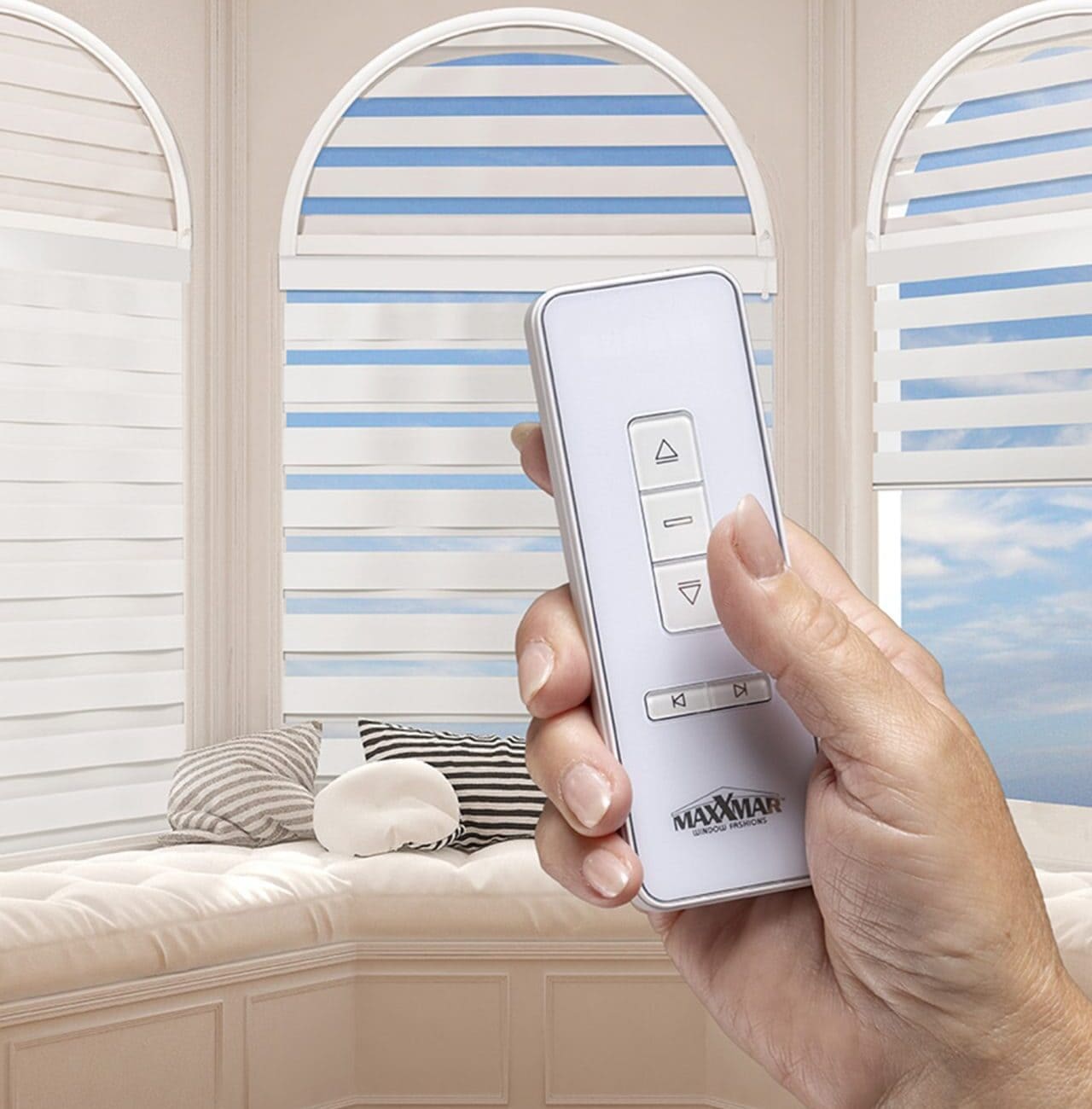 Hand holding remote control for Maxxmar motorized dual shades in a bright room with large arched windows.
