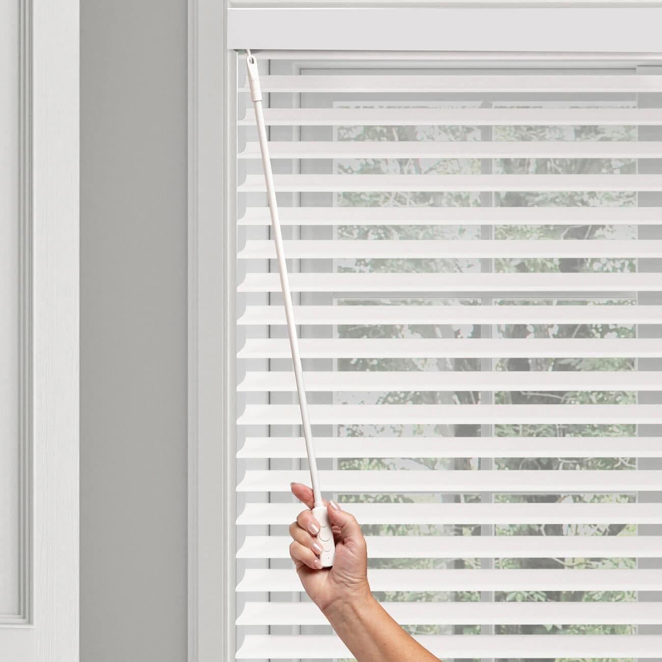 Person adjusting white horizontal window blinds with a wand, letting in natural light, in a modern home interior.
