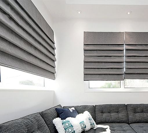 Contemporary living room featuring two large windows with gray fabric Roman blinds, a dark gray sectional sofa, and decorative pillows.
