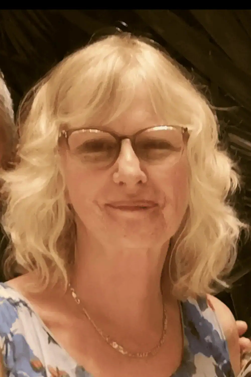 Close-up of a woman with blonde curly hair, glasses, and a floral top, smiling at the camera.