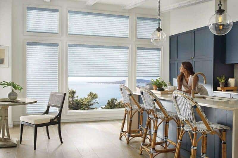 Modern kitchen with large windows overlooking the ocean, featuring a woman sitting at the counter, bar stools, and contemporary decor.