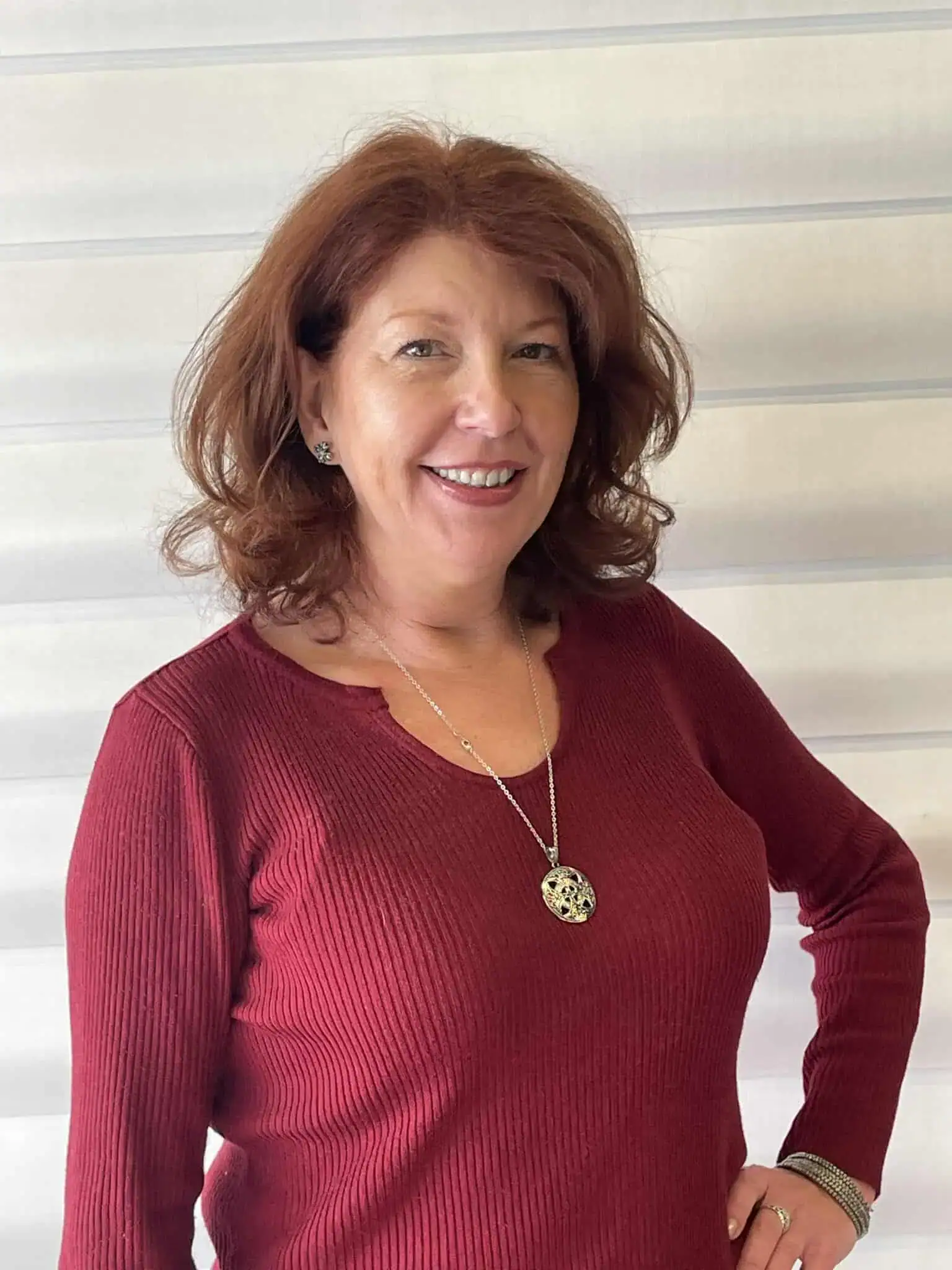 Woman with red hair smiling, wearing a red sweater and jewelry, standing against a light background.